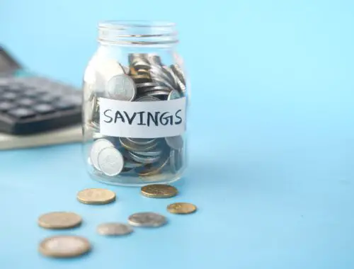The benefits of saving money are shown in this picture by having coins in a clear glass jar marked savings on a blue background